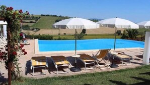 Zona de piscina con sillas de descanso y sombrillas, rodeada de un jardín ajardinado y vistas al campo.