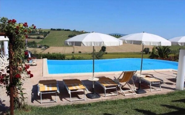 Zona de piscina con sillas de descanso y sombrillas, rodeada de un jardín ajardinado y vistas al campo.