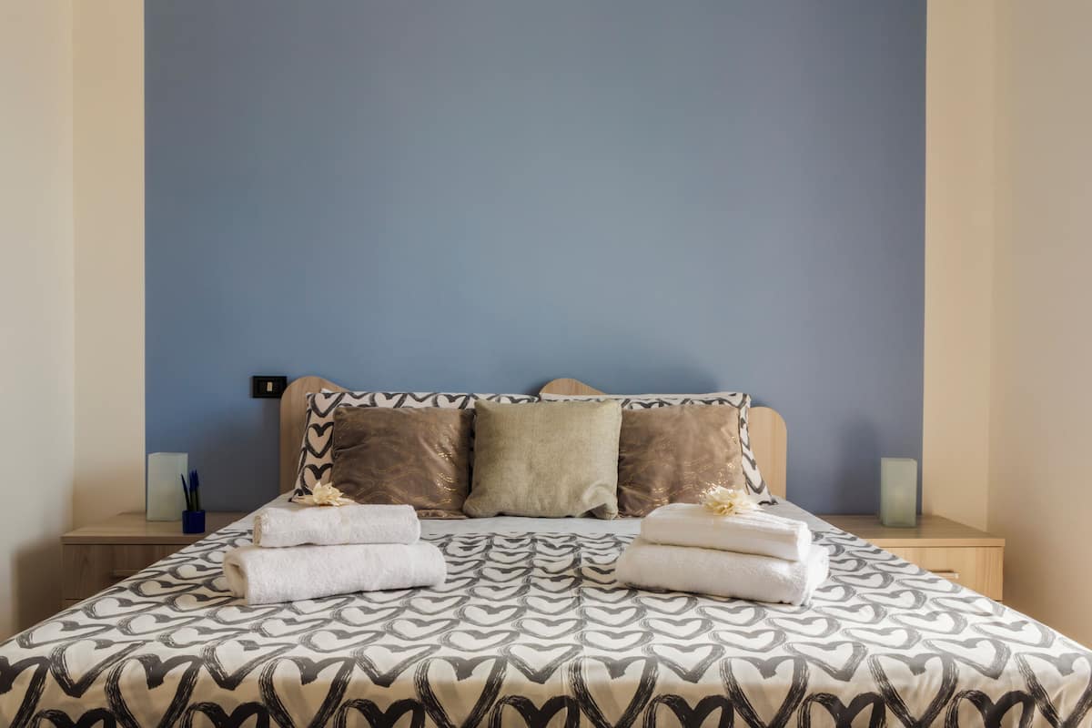 A neatly made bed with a patterned bedspread, two pillows, and folded towels on a wooden nightstand.