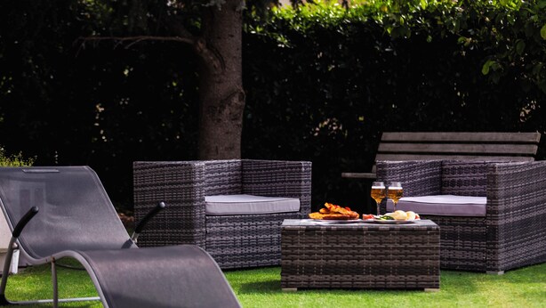 Un espace patio aménagé avec des meubles en osier, notamment un canapé et un fauteuil, ainsi qu’une petite table sur laquelle se trouvent une assiette de nourriture et des boissons.