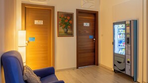Une chambre d’hôtel avec deux portes en bois, un fauteuil bleu, une lampe fixée au mur, un tableau représentant des fleurs et un distributeur automatique.