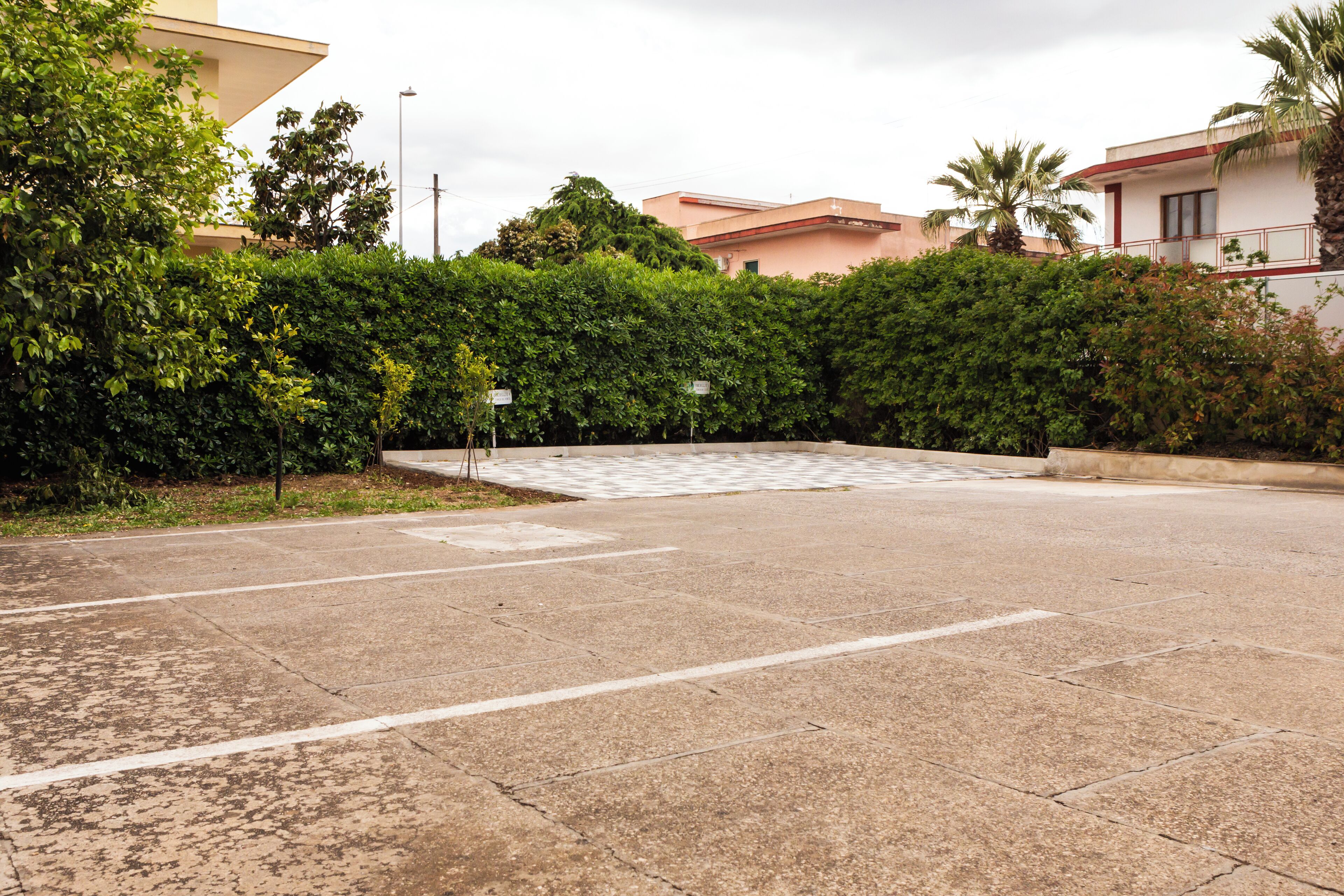 A parking area with a concrete surface, surrounded by dense greenery and a few buildings in the background.