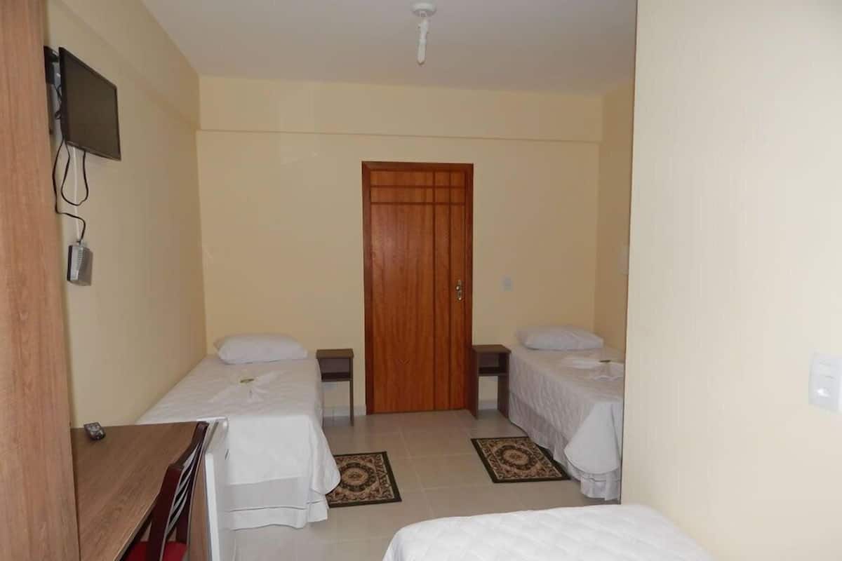 Une petite chambre propre, avec deux lits simples, une porte en bois, un téléviseur fixé au mur et une petite table avec une chaise.