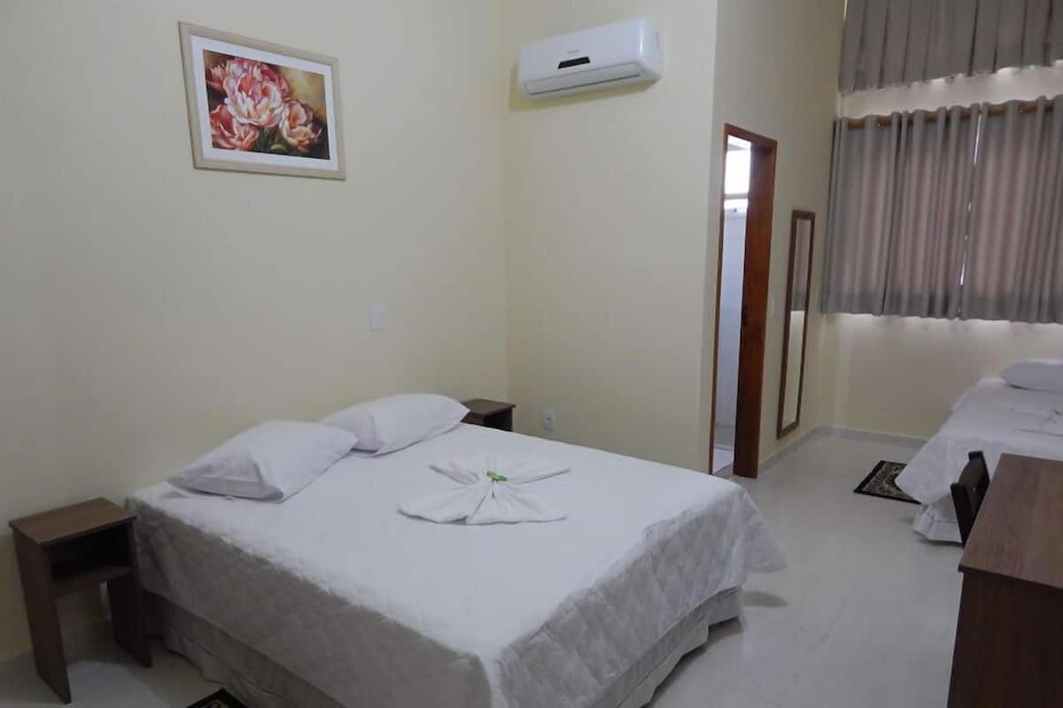Une chambre d’hôtel avec un lit, une table de chevet, une petite table, un appareil de climatisation, un tableau encadré au mur et une fenêtre avec des rideaux.