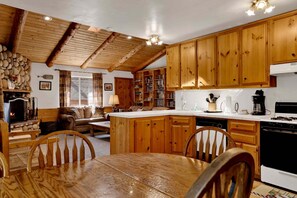 A rustic kitchen with wooden cabinets, a stone fireplace, and a dining area with a wooden table and chairs. - Forest Treehouse # 1473 (Big Bear Lake)