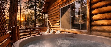 A log cabin with a hot tub, wooden deck, and a large window overlooking a forest.