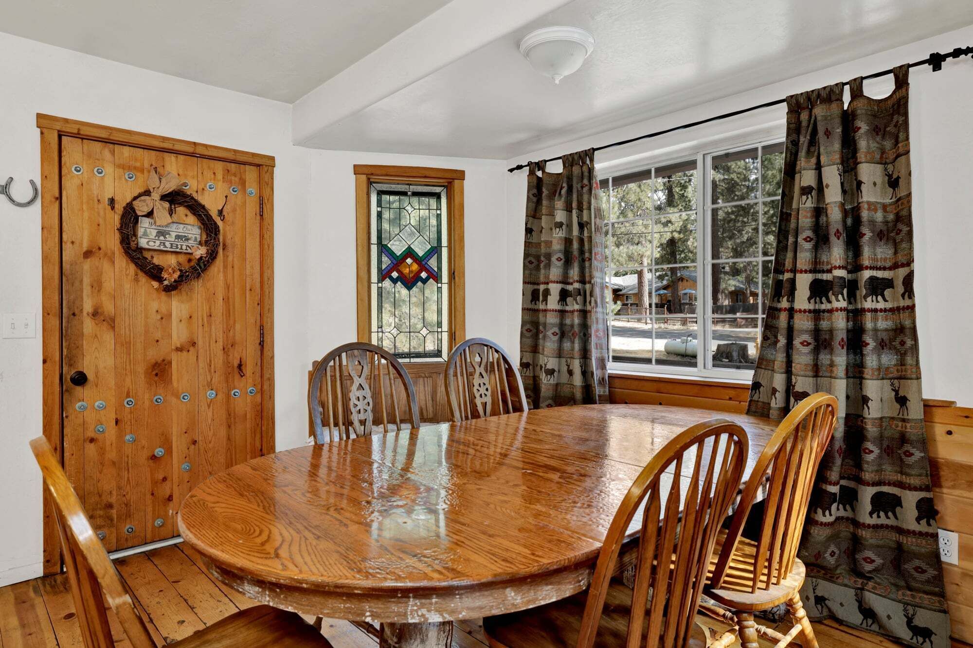 A wooden dining table with four chairs, a wooden door with a wreath, a stained glass window, and a view through a window with patterned curtains.