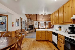 A cozy kitchen with wooden cabinets, a stone fireplace, and a dining area with a wooden table and chairs. - Forest Treehouse # 1473 (Big Bear Lake)