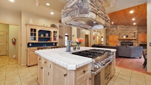 A spacious kitchen with a central island, wooden cabinetry, a stone accent wall, and a fireplace. - Four Happy Bears Estate #1277 (Big Bear Lake)