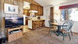A cozy room with a wooden wall, a TV, a small kitchen area, and a dining table with chairs. - Cedar Pines #1529 (Big Bear Lake)