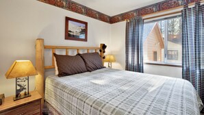 A bedroom with a wooden headboard, a bed with a plaid bedspread, two bedside lamps, a framed picture on the wall, and a window with curtains. - Cedar Pines #1529 (Big Bear Lake)