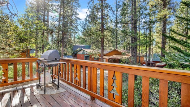 A wooden deck with a barbecue grill, surrounded by tall pine trees and small cabins. - Cedar Pines #1529 (Big Bear Lake)