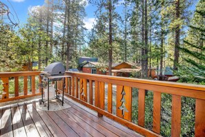 A wooden deck with a barbecue grill, surrounded by tall pine trees and small cabins. - Cedar Pines #1529 (Big Bear Lake)