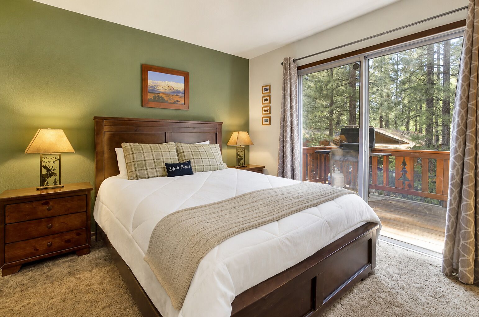 A bedroom with a large bed, two nightstands, a window with a view of trees, and a wooden deck outside.