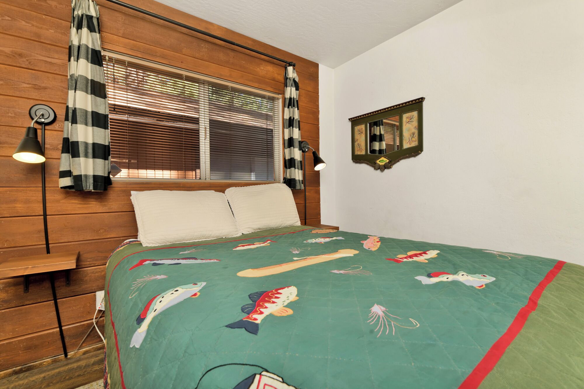 A bedroom with a wooden headboard, a bed with a green quilt featuring fish and sea creatures, a window with blinds, and a wall-mounted mirror.