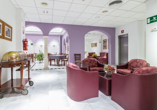 Un vestíbulo con piso de mármol, sillones tapizados en rojo, un carrito con una cúpula y una mesa con sillas.