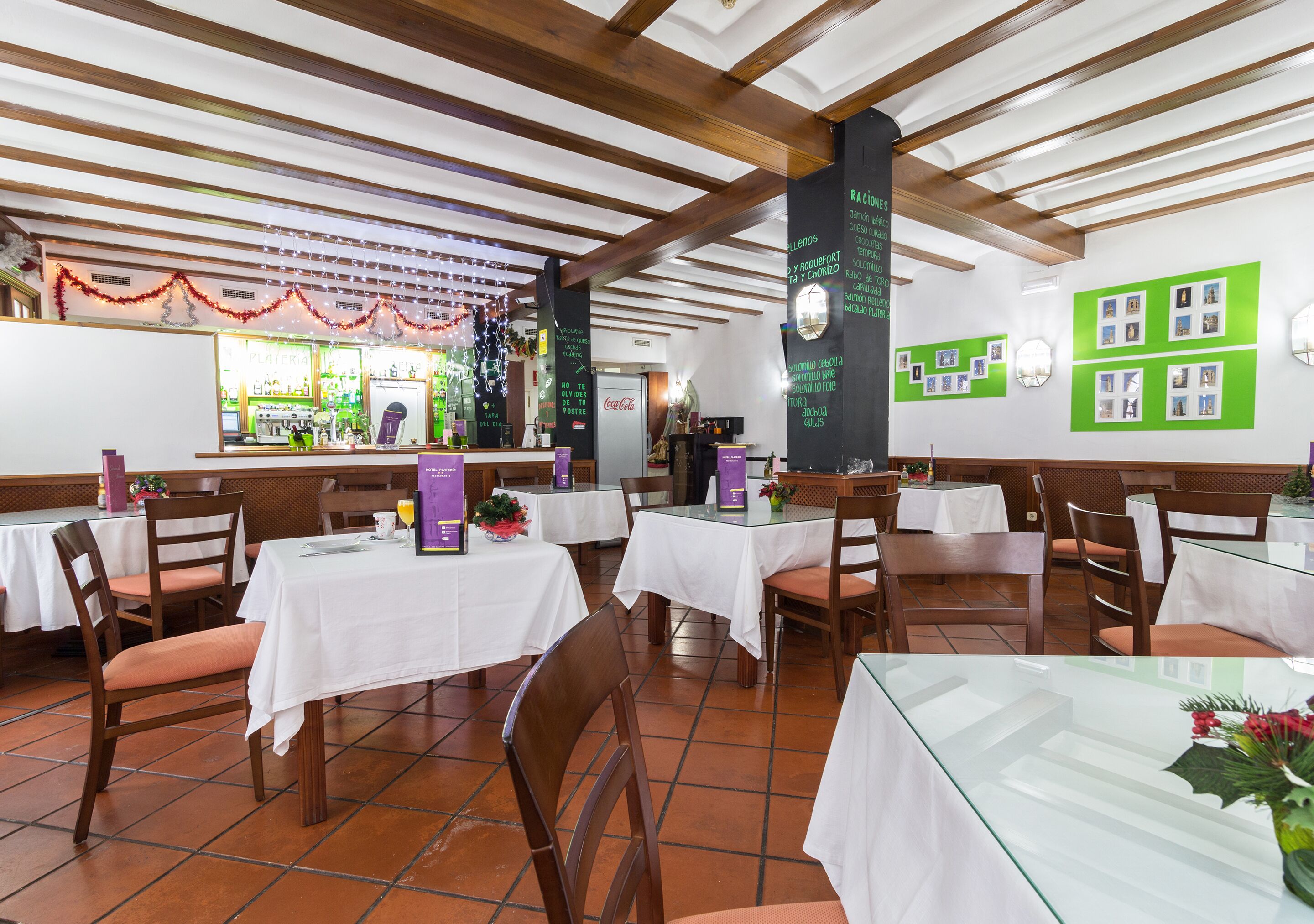 A restaurant with white tablecloth-covered tables, wooden chairs, and a tiled floor. There are green bulletin boards on the walls and a Coca-Cola refrigerator.