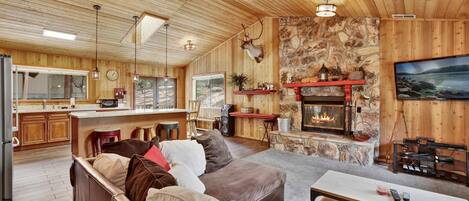 A cozy cabin interior with a stone fireplace, wooden walls, a kitchen area, and a seating area with a sofa and coffee table.
