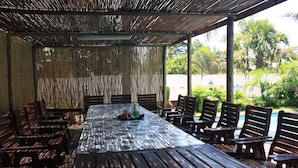 A covered outdoor dining area with a long wooden table and benches, surrounded by a bamboo fence, overlooking a pool and greenery. - Big Ben Lodge (Polokwane)