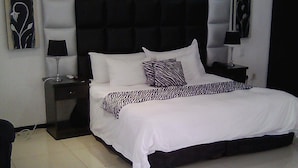A bedroom with a large bed, a headboard with a tufted design, two bedside tables with lamps, and artwork on the walls. - Big Ben Lodge (Polokwane)