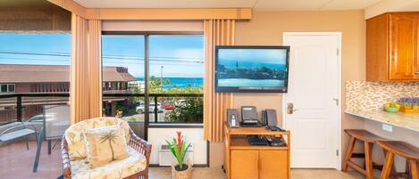 A modern kitchen with a large TV, a dining area with a wicker chair, and a view of the ocean.