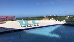 A rooftop pool with three blue lounge chairs and a view of the ocean and buildings in the background. - Blue Residences at Blue Mall (Lowlands)