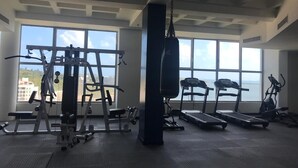 A gym with cardio machines, weightlifting equipment, and a punching bag, with large windows offering a view of the city. - Blue Residences at Blue Mall (Lowlands)