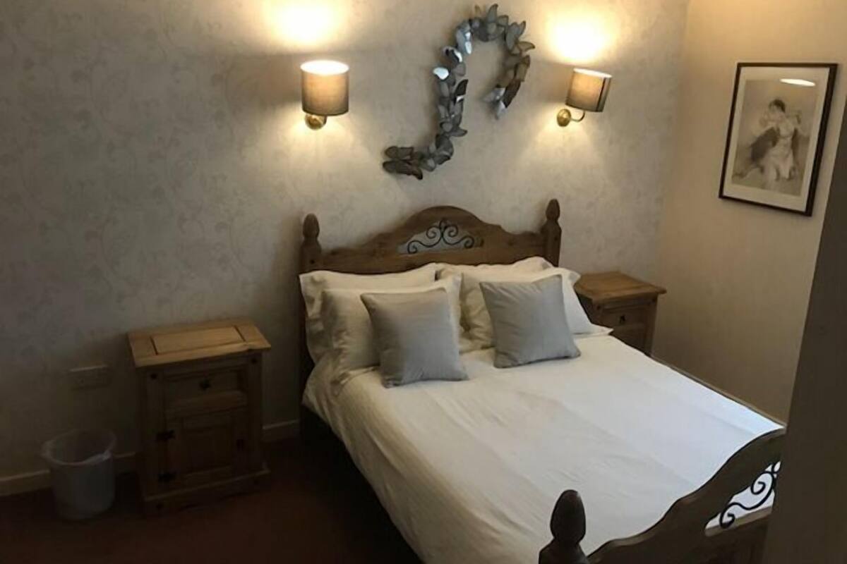 Ein Schlafzimmer mit einem hölzernen Kopfende, zwei Nachttischen, einem Beistelltisch, einem Mülleimer und einem gerahmten Bild an der Wand.