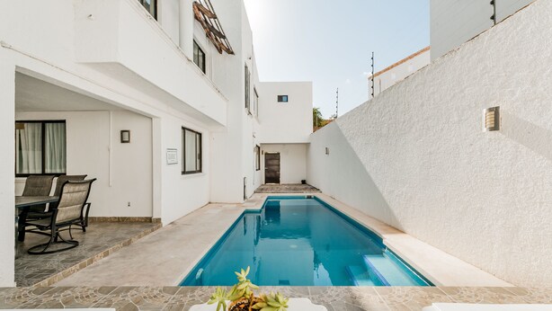 Un espace piscine moderne avec une piscine à l’eau d’un bleu limpide, des murs blancs et un coin repas comprenant une table et des chaises.