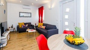 Ein modernes Wohnzimmer mit einer Couch, roten Sesseln, einem Esstisch mit Obst und einem Fernseher.