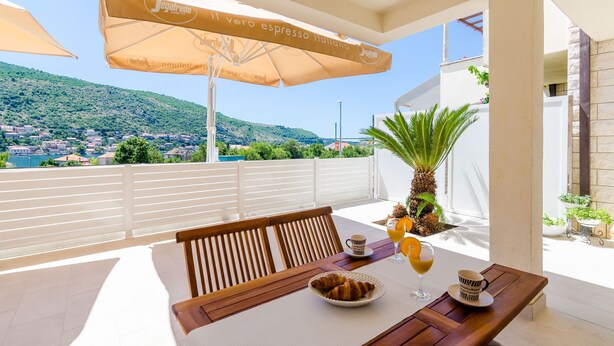 Eine Terrasse mit Tisch, Stühlen, einer Schüssel Croissants, einem Glas Orangensaft und einer Tasse. Sonnenschirme und im Hintergrund eine Ansicht einer Stadt und Hügel.