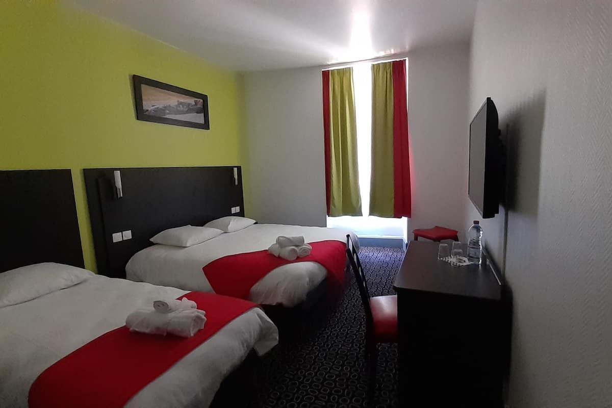 A hotel room with three beds, a small table with a bottle and glasses, a TV on the wall, and a window with curtains.