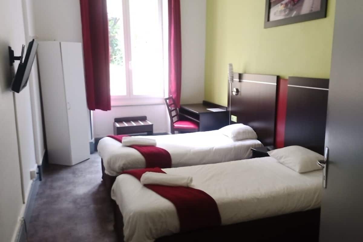 A hotel room with two single beds, a desk, a chair, a wardrobe, and a window with curtains.