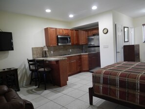 A compact kitchen with wooden cabinets, a microwave, and a refrigerator. There is a small dining area with a chair and a table. The room has a bed with a plaid blanket and a television mounted on the wall. - Shell Studios Hallandale Beach (Hallandale Beach)