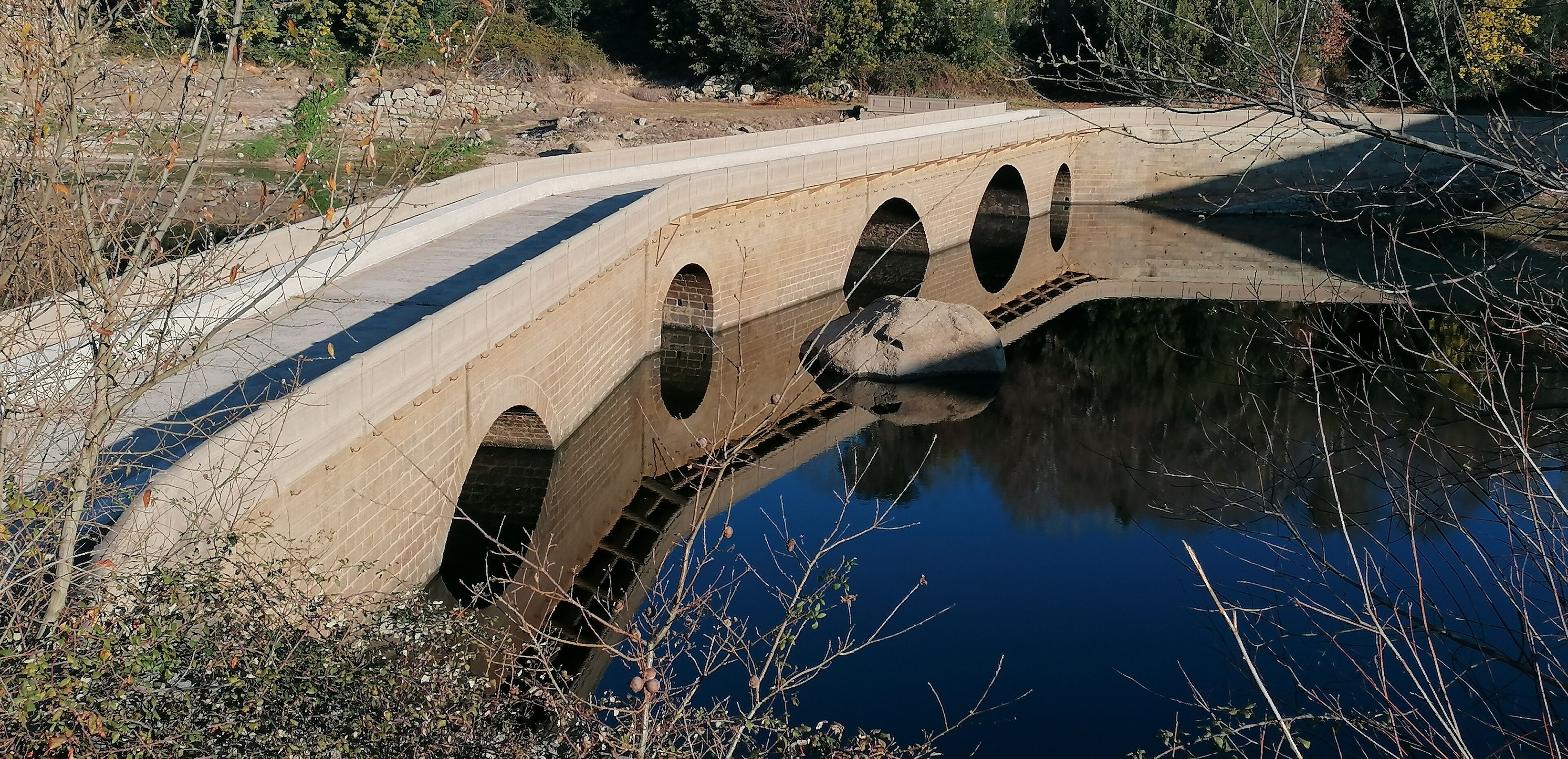 A stone bridge over a calm body of water with arched openings, surrounded by natural vegetation.
