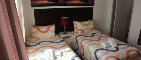 A hotel room with two beds, each with a patterned bedspread, a nightstand with a lamp, and a wall-mounted artwork depicting a boat on water.