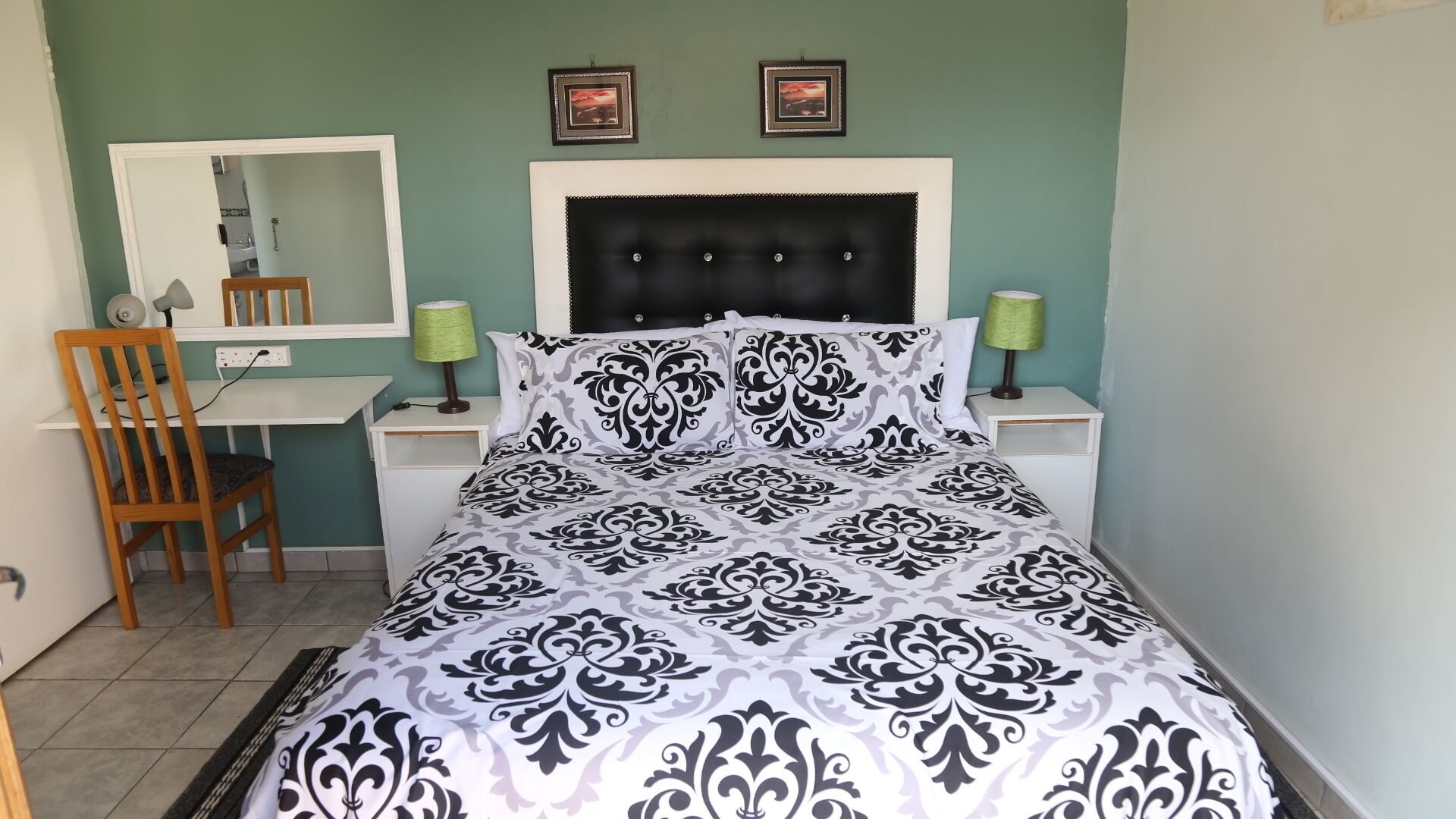 Um quarto com cama, escrivaninha, cadeira, espelho e duas molduras na parede.