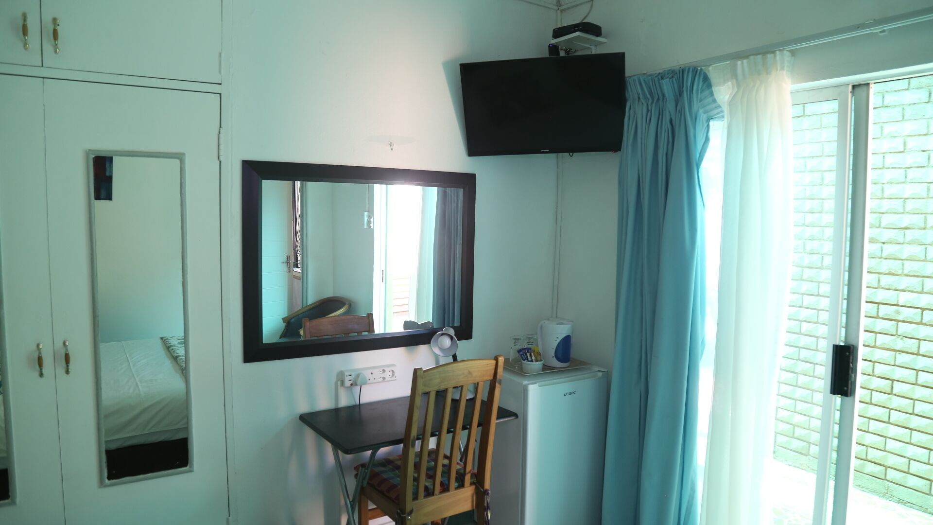 A room with a desk, chair, mirror, television, and a window with curtains.