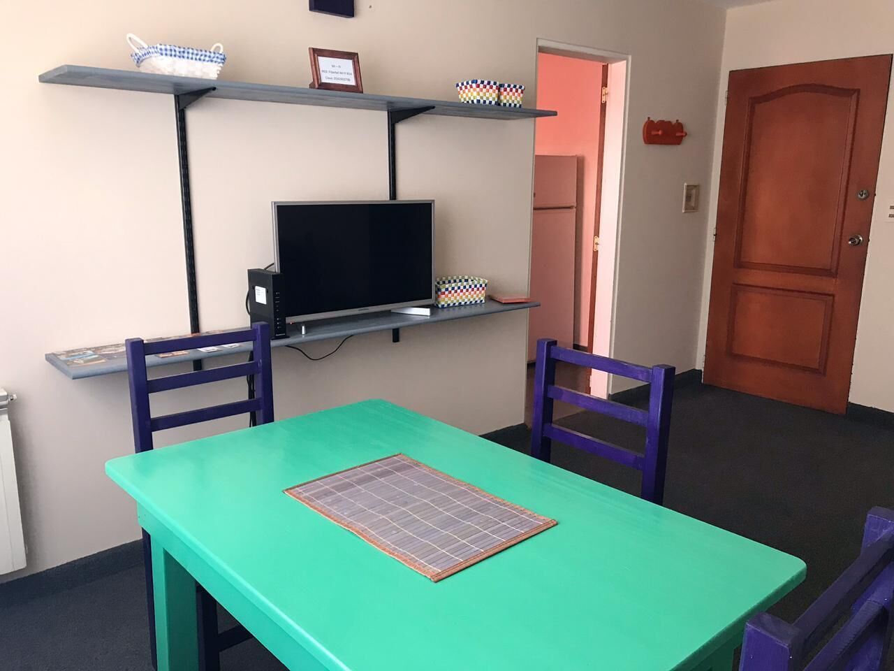 A room with a green table, two purple chairs, a TV on a shelf, and a door in the background.