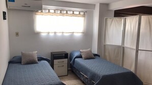 Una habitación pequeña y limpia con dos camas individuales, una mesita de noche, una ventana con cortinas e un equipo de aire acondicionado.