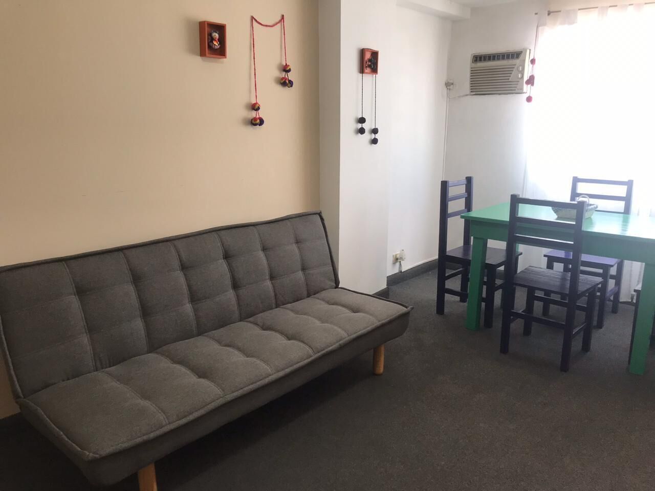 A room with a grey sofa, a green table, and two wooden chairs. There are wall decorations and an air conditioning unit.