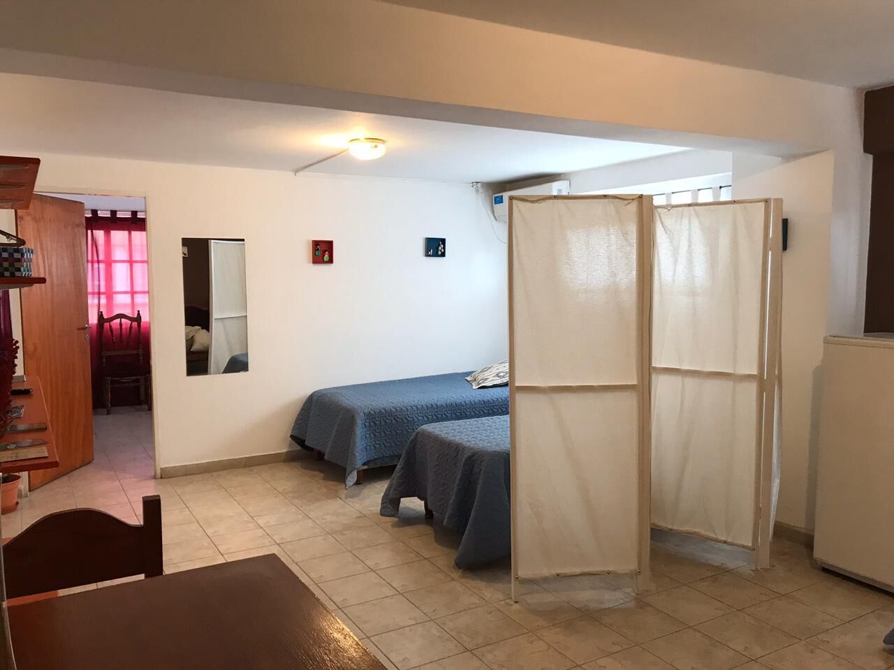 A small, well-lit room with two beds, a wooden chair, a table, and a kitchenette.