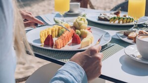 Une table à manger installée sur la plage, avec des assiettes de fruits et des verres de jus, des palmiers et l’océan en arrière-plan.