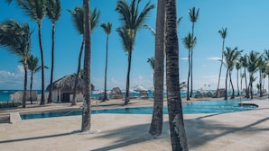 Un espace piscine agrémenté de palmiers et de cabanes de plage, avec vue sur l’océan.