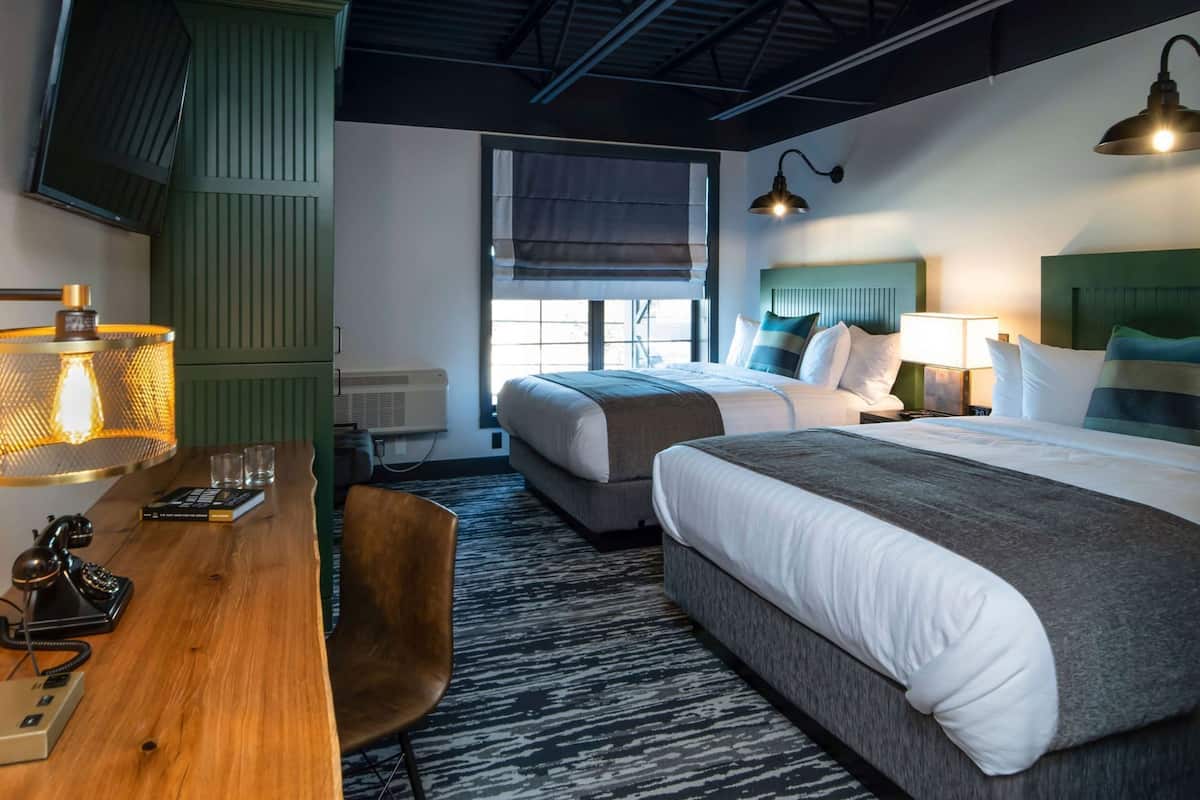 A hotel room with two beds, a desk with a lamp, a chair, and a window with blinds.