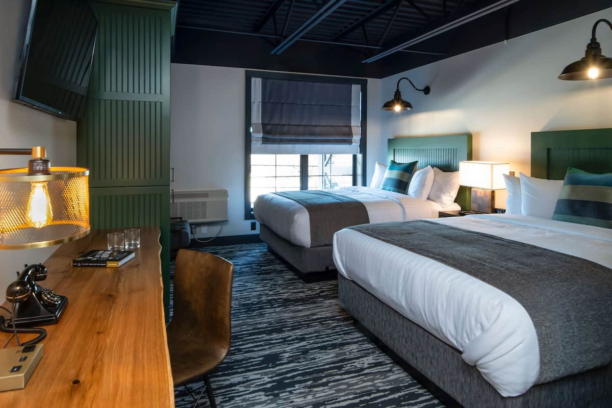 A hotel room with two beds, a wooden desk with a telephone, a chair, and a window with a view.
