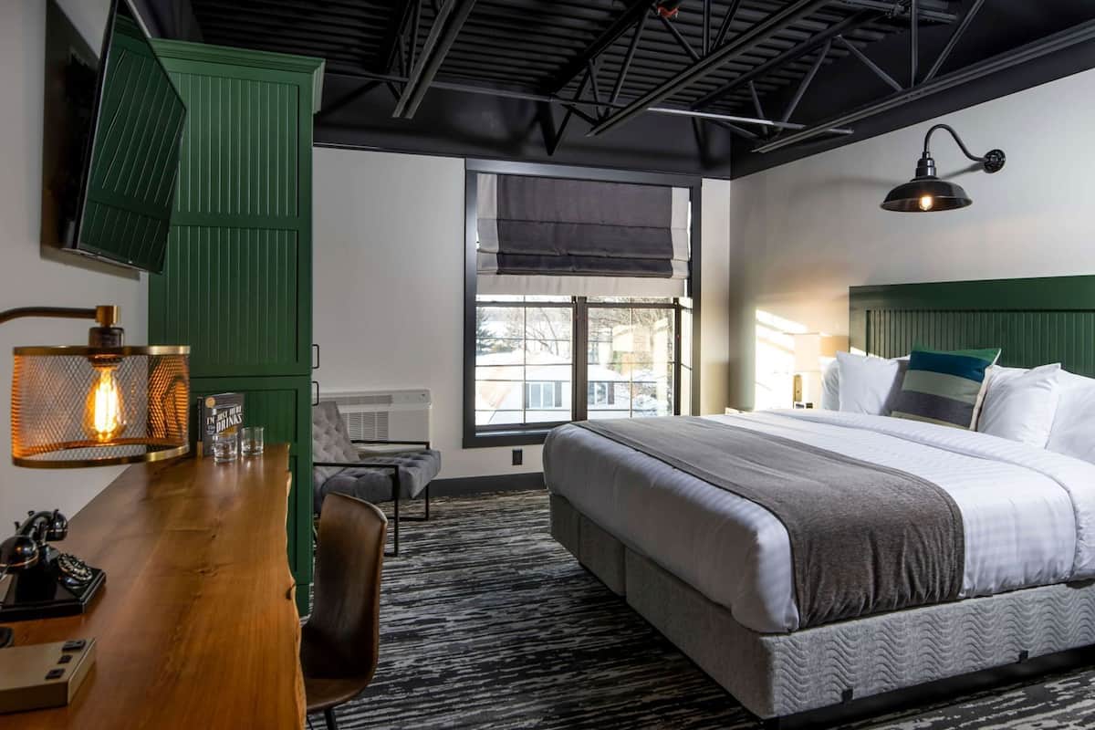 A hotel room with a bed, a desk with a lamp, a chair, a window with a view, and a ceiling with exposed beams.
