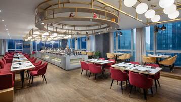 A modern restaurant with round tables and red chairs, a central bar area, and large windows offering city views.