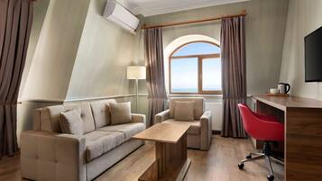 A modern hotel room with a sofa, a wooden coffee table, a red office chair, a desk, and a large window with curtains.