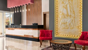 A reception area with a reception desk, two red chairs, a round table, and a decorative wall with a relief sculpture. - Ramada by Wyndham Van (Edremit)